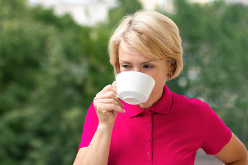 Middle-aged mature senior woman is drinking tea or coffee outdoor in a sunny summer day in a pink or red polo shirt. Thoughtful female pensioner holding a mug. Relaxing and thinking, happy retirement.