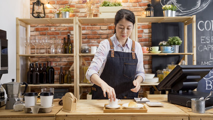 elegant young girl barista putting cup of hot cappuccino down on tray with croissant in wooden counter bar. woman waitress working in coffeehouse preparing customer order. modern coffee shop.
