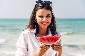 Portrait of smiling woman relaxing holding and eating a slice of watermelon on the tropical beach.Summer vacations