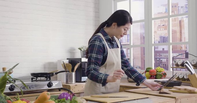 slow motion of happy young chinese woman in apron cooking at home. lunch preparation at modern kitchen. asian housewife checking recipe on internet in mobile touchpad with fresh vegetables on table