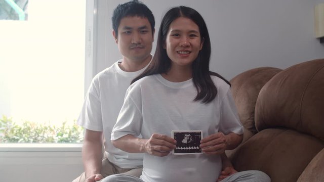 Young Asian Pregnant couple show and looking ultrasound photo baby in belly. Mom and Dad feeling happy smiling peaceful while take care child lying on sofa in living room at home concept. Slow motion.
