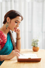 Pretty young Indian woman in traditional sari dress sitting at desk at home and checking plans in her diary