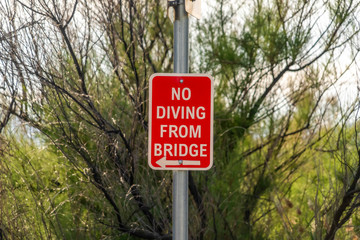 No Diving From Bridge sign with arrow against trees and sky on a sunny day