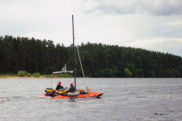 Obraz premium An orange inflatable sailing catamaran with a lowered sail floats under the outboard motor on the water against the background of the far green shore on a bad weather cloudy summer day, water tourism