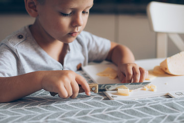 Cute boy 4-5 years old with knife cutting a cheese on the cutting board on the table in the...