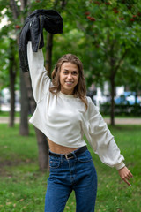 Happy Caucasian girl. Young beautiful European long free hair walks in city park in cheerful mood. Smile, white teeth. Wearing pullover. In hands black leather jacket. To enjoy life. Lifestyle