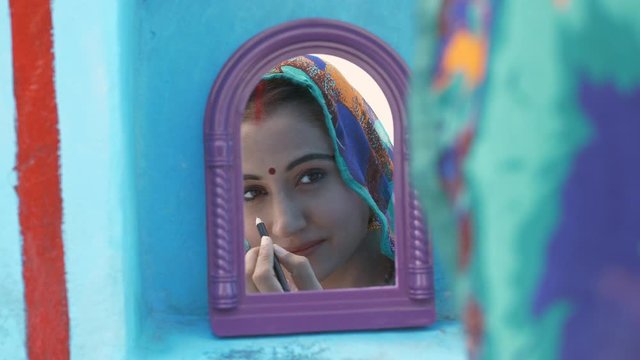 Close up shot of a young woman getting ready to go for work - village life in India. Beautiful Indian lady applying kajal looking in the small wall mirror  getting ready to go out on the field work...