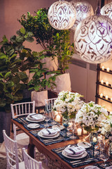 Christmas table with white flowers, blue designer plates, glasses and white with white chairs and a candle mural.