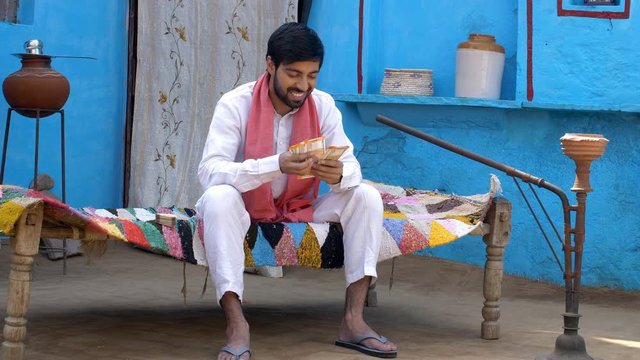 Indian villager happily counting his monthly earnings - financial concept  village home. Attractive young man counting his bundle of Indian rupee notes while relaxing on a charpai - monthly income