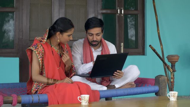 Young educated villager teaching basic usage of computer to his wife - village lifestyle. Beautiful Indian wife learning usage of computer from her husband while sitting on a charpai - relationship...