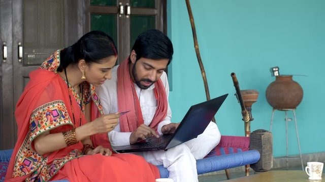 Indian village couple doing card payment using laptop - online shopping concept. Young beautiful wife holding a debit/credit card in her hand and telling her husband card details to do online payment