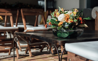 Bouquet of orange and white flowers in a glass vase on a black metal table on a carpet and wooden chairs. Decoration for social events.
