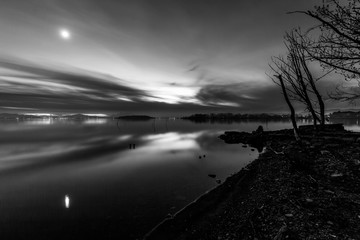 Moon above a lake with its reflection on the Trasimeno lake surface at dusk and trees on the shore
