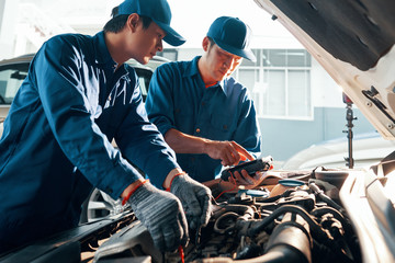 Serious mechanic showing screen of multimeter to his coworker when they are measuring car battery voltage in garage