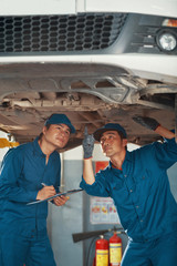 Mechanic pointing at bottom of suspended car and explaining problems to his coworker taking notes...