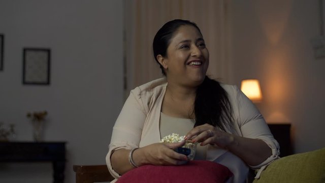 Overweight female taking a handful of popcorn and eating in front of the TV at home - Sedentary and unhealthy lifestyle. Chubby woman enjoying a silly programme on television and laughing while eat...