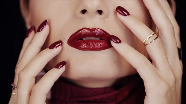 Closeup of female face with red lipstick and hands with red manicure. Golden rings on female fingers. She touches her face with her fingertips. Passion and temptation. In studio on black background.