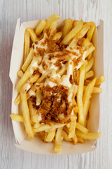 Fastfood: french fries with cheese sauce and fried onion in a paper box on a white wooden background, top view. Flat lay, from above, overhead. Close-up.