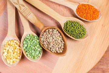 Various uncooked legumes in wooden spoons, top view close-up