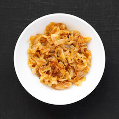 Homemade caramelized onions on a white plate on a black surface, top view. From above, overhead, flat lay.