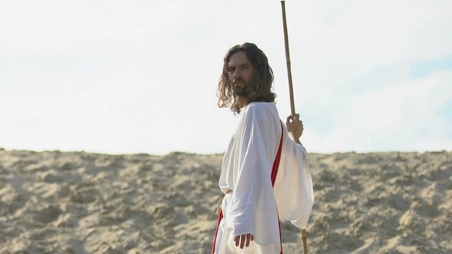 Jesus with staff in robe and sash walking on sandy hill, stretching helping hand