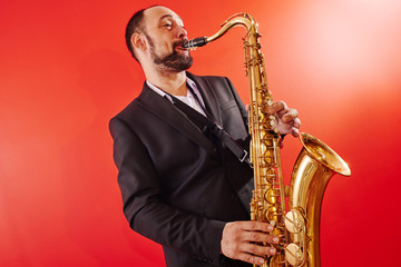 Plakat Portrait of professional musician saxophonist man in suit plays jazz music on saxophone, red background in a photo studio
