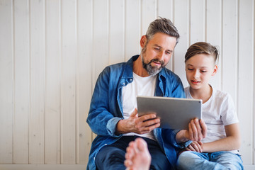 Mature father with small son sitting indoors, using tablet. Copy space.