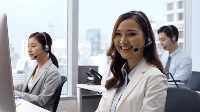 Young asian woman wearing microphone headset working in customer service support help desk with colleagues looking to camera and smiling