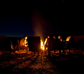 The group of friends are sitting near the bonfire in the night and talking about something
