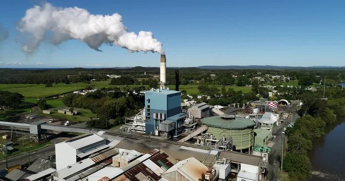 Sugar mill processing sugar cane to sweet product in rural NSW on shores of a river with tall chimney exhausting steam to blue sky.