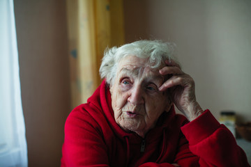 Lonely old woman in her house, sitting at a table in a red jacket. The plight of pensioners.