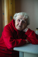 A very old woman in her house, a portrait sitting at a table in a red jacket. The plight of pensioners.