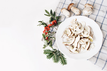 Autumn food arrangement. Composition of whole and sliced porcino mushrooms, ceps on plate. Rowan berries, leaves, fir branches and checkered tea towel on white table background. Fall design, flat lay