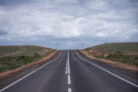Steppe panorama of a desert landscape summer day. An asphalt road with markings goes forward to the horizon. Traveling among the sands and shrubs. Background image. Dull sky with clouds.