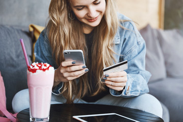 Good-looking caucasian girl with fair hair, sitting near coffee table, drinking cocktail, holding smartphone and credit card, paying for drink while relaxing, watching funny videos in digital tablet