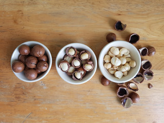 Unpeeled, semi peeled, and peeled macadamia nuts in white ceramic bowls on a wooden table 