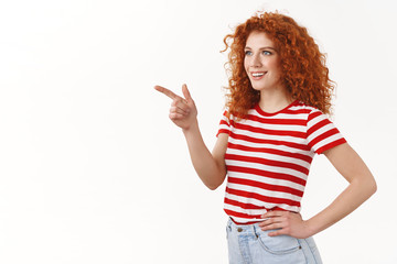 Interesting what is it. Intrigued charismatic half-turned redhead curly woman check out cool offer pointing looking left smiling impressed amused feel uplifted show shop assistant picked product