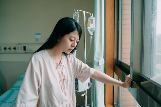 Young Depressed Asian Korean Woman Standing By Bed And Leaning Against Window With One Hand In Hospital Worried About Her Illness. Medicine And Health Care Concept. Sad Girl Patient In Hospital Gown