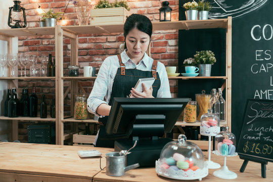 asian chinese woman rendering account with access point of sale terminal machine in coffee shop. elegant female waitress in apron holding note of customer order and touching on cash register in cafe