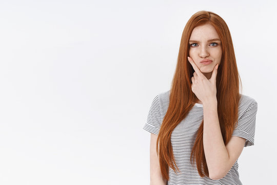 Curious doubtful young perplexed redhead girlfriend looks suspicious rubbing chin smirking displeased frowning thinking, pondering evidences standing hesitant white background