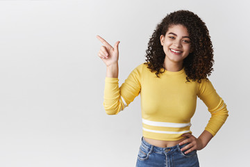 Carefree confident friendly smiling upbeat lively girl curly hairstyle pointing upper left corner hold hand waist self-assured relaxed grinning delighted show awesome copy space offering goods