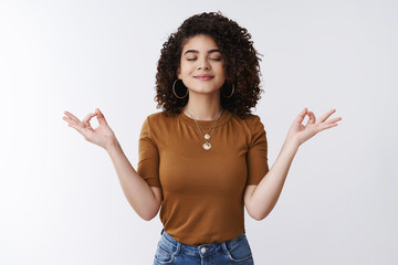 Om girl feels peace patience. Attractive carefree relaxed happy young woman curly shirt hairstyle close eyes smiling delighted meditating hands sideways nirvana lotus pose, breathing practice yoga - obrazy, fototapety, plakaty