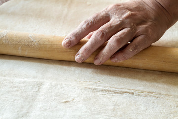 Senior woman hands are rolling out dough in flour with rolling pin in her home kitchen. Homemade noodle or pasta production by grandma. Closeup, selective focus