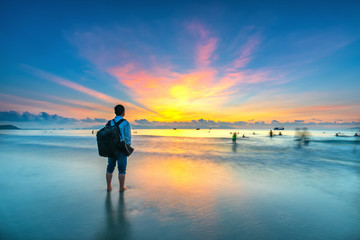 Fototapeta premium Silhouette of man backpacker on the beach looking at magical dramatic sunrise. The man standing on the sandy beach