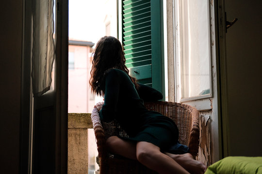 Woman In Tight Knit Dress Sitting In Wicker Chair On A Balcony With Green Wooden Shutters In Traditional Italian Hotel. Female Solo Travel Concept