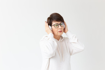 Cute positive middle-aged woman in sweater and glasses is listening to music with wire headphones standing on a white background. Concept of hobbies and subscriptions to favorite radio station.