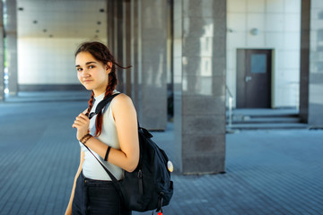 Young beautiful student with backpack on her shoulder goes to school, close-up. Schoolgirl with two braids standing in front of college, blurred background. September 1, the beginning of school year.