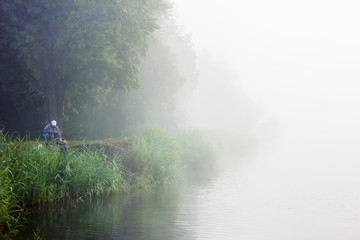 Fisherman on river bank. Elderly man with fishing rod in white morning fog. Summer rural landscape with green trees and reeds