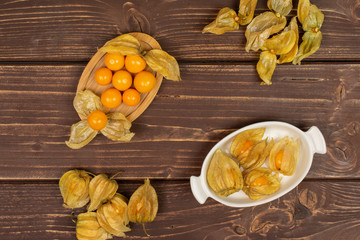 Lot of whole lot of pieces of fresh orange physalis on oval bamboo coaster in white oval ceramic bowl flatlay on brown wood