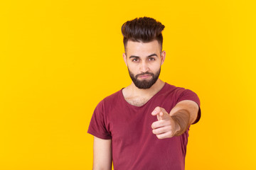 Fototapeta premium Cute young hipster man with a beard pointing at the camera is posing on a yellow background with copy space. Concept of choice and win.